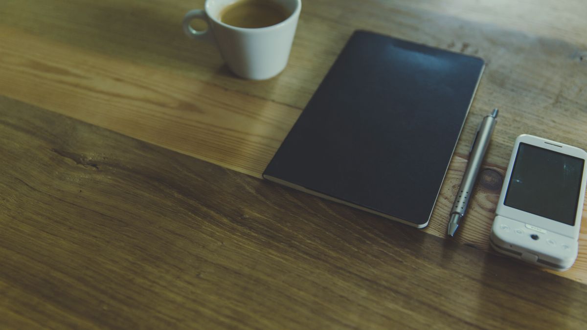 A white coffee mug beside a black notebook and a phone on a wooden table